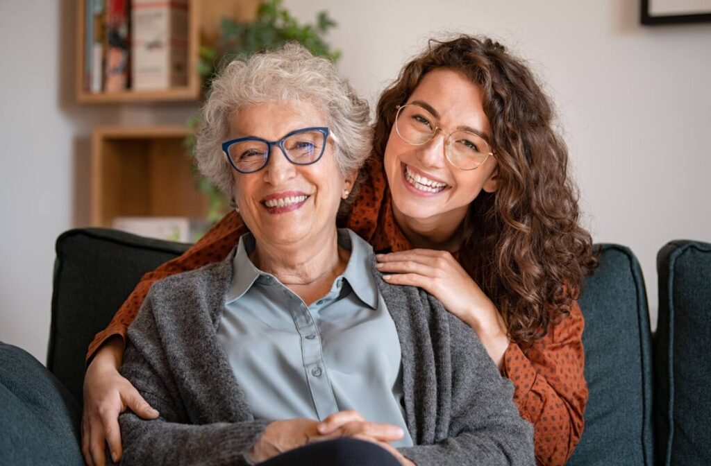 A smiling senior woman with short grey hair wearing bold blue rectangular eyeglasses sits on a couch next to a younger woman with curly hair wearing round gold metal frames.